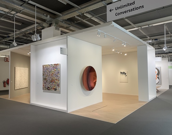 art exhibition booth with paintings on white walls inside big event venue, grey carpet flooring